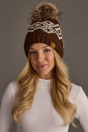 A woman with long blonde hair models the Panache Apparel Collection’s Brown & White Patterned Pom Hat, featuring a white zigzag design and a fur pom-pom on top. She pairs it with a white ribbed turtleneck and stands against a plain gray background.