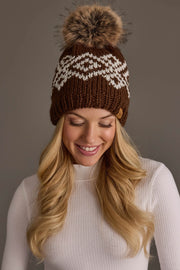 A woman with long blonde hair smiles and looks down while wearing the Panache Apparel Collection’s Brown & White Patterned Pom Hat, a brown knit beanie featuring a white geometric design and pom-pom, paired with a white ribbed turtleneck shirt against a neutral background.