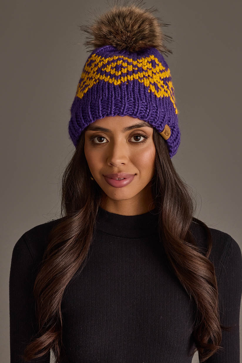 A woman with long brown hair is wearing the Panache Apparel Collection Purple & Gold Patterned Pom Hat, featuring yellow zigzag accents and a brown faux fur pom-pom, paired with a black ribbed turtleneck sweater against a gray background.