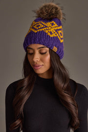 A woman with long dark hair, wearing the Panache Apparel Collection’s Purple & Gold Patterned Pom Hat featuring a yellow design and fur pom-pom pairs it with a black top as she looks down with a neutral expression.