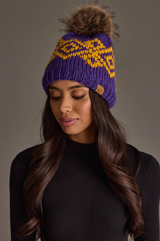 A woman with long dark hair, wearing the Panache Apparel Collection’s Purple & Gold Patterned Pom Hat featuring a yellow design and fur pom-pom pairs it with a black top as she looks down with a neutral expression.