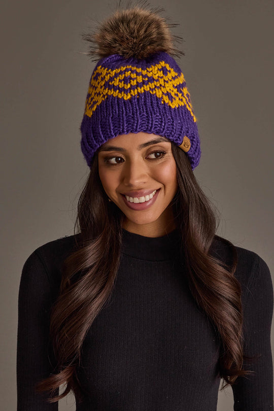 A woman smiles while wearing the Panache Apparel Collection’s Purple & Gold Patterned Pom Hat featuring a furry pom-pom paired with a black long-sleeve top against a neutral background.