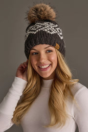 A smiling woman with long blonde hair wears the Panache Apparel Collection’s Dark Gray & White Patterned Pom Hat, featuring a large fur pom-pom, paired with a white ribbed long-sleeve shirt, as she stands against a plain gray background.