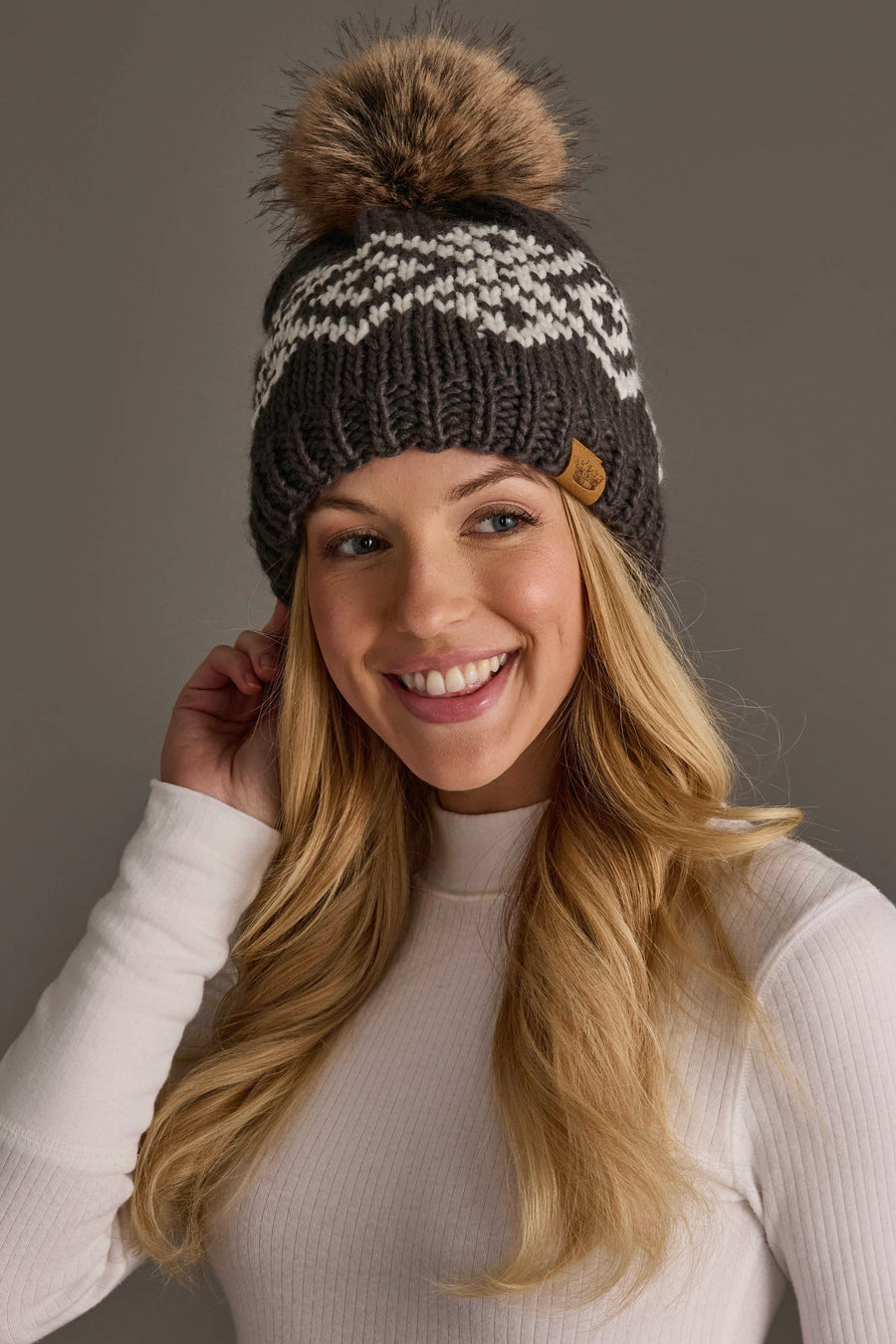 A smiling woman with long blonde hair wears the Panache Apparel Collection’s Dark Gray & White Patterned Pom Hat, featuring a large fur pom-pom, paired with a white ribbed long-sleeve shirt, as she stands against a plain gray background.