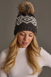 A woman with long blonde hair models the Panache Apparel Collection’s Dark Gray & White Patterned Pom Hat, featuring a bold zigzag design and a large brown pom-pom, paired with a white ribbed top against a plain gray background.
