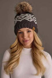 A woman with long blonde hair wears the Panache Apparel Collection’s Dark Gray & White Patterned Pom Hat, featuring a large fur pom-pom. She is dressed in a light ribbed sweater and stands against a neutral background.