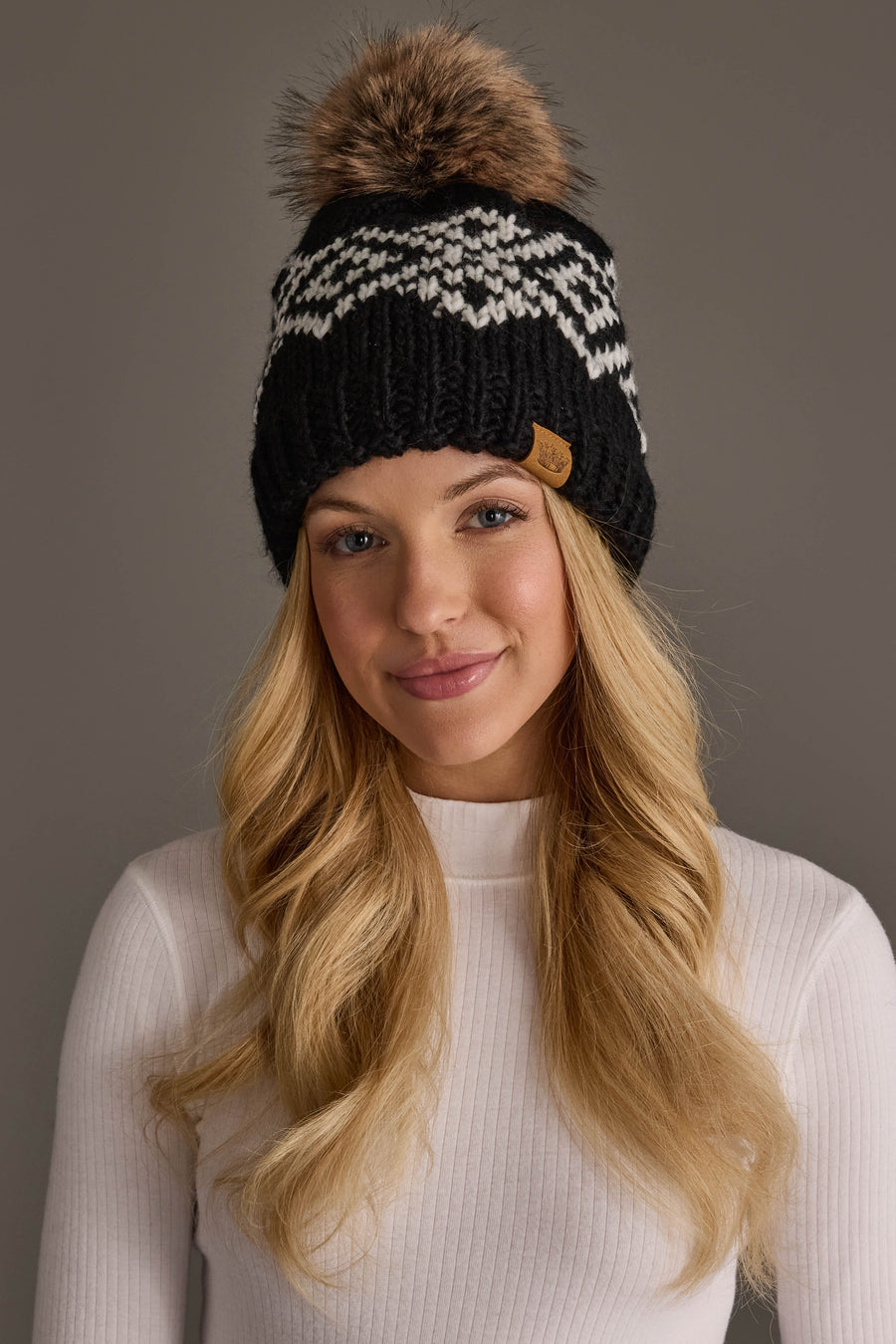 A woman with long blonde hair wears a white ribbed sweater and the Panache Apparel Collection Black & White Patterned Pom Hat, smiling softly against a neutral background.