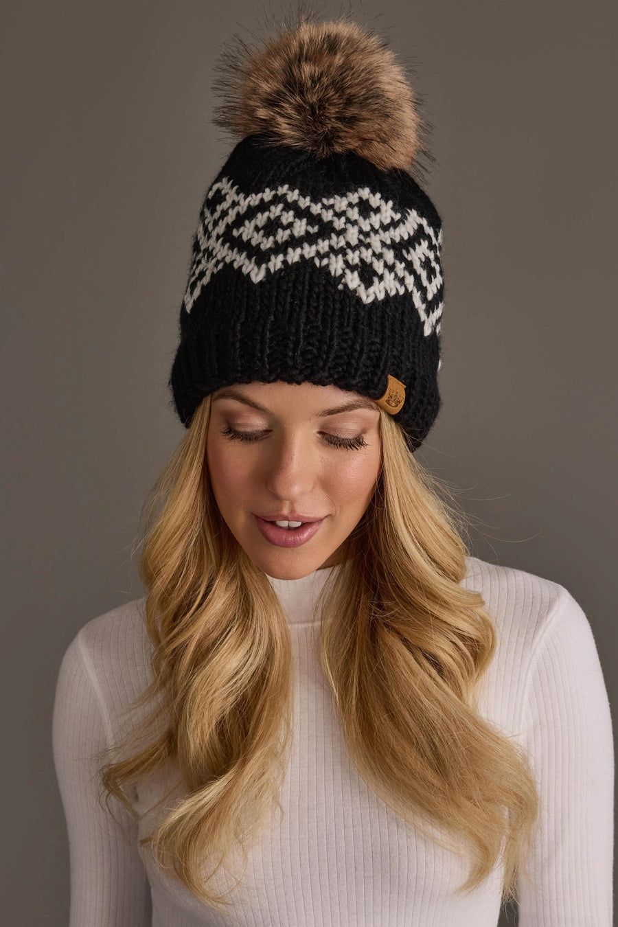 A woman with long blonde hair wears the Panache Apparel Collection Black & White Patterned Pom Hat, featuring a white geometric design and a large brown faux fur pom-pom, along with a white ribbed sweater, as she looks downward against a plain gray background.