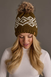 A woman with long blonde hair looks down and smiles while wearing the Panache Apparel Collection’s Olive & White Patterned Pom Hat, a green knit beanie featuring a white pattern and a fur pom-pom, paired with a white ribbed sweater, set against a neutral background.