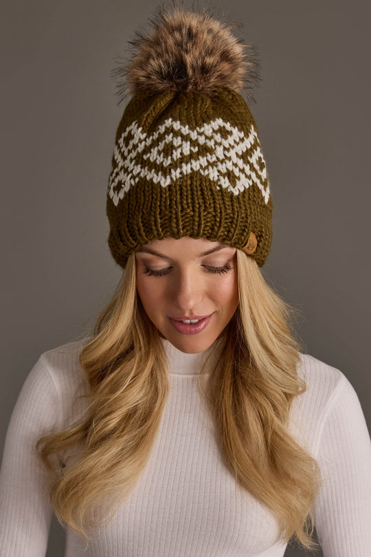 A woman with long blonde hair looks down and smiles while wearing the Panache Apparel Collection’s Olive & White Patterned Pom Hat, a green knit beanie featuring a white pattern and a fur pom-pom, paired with a white ribbed sweater, set against a neutral background.