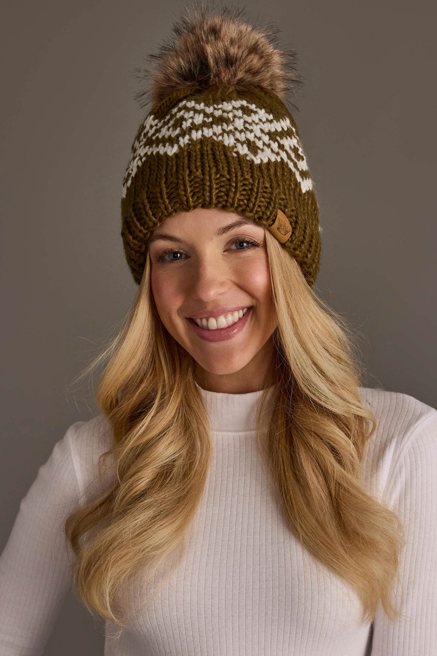 A smiling woman with long blonde hair wears a white sweater and the Panache Apparel Collection’s Olive & White Patterned Pom Hat, featuring a green knit design with a white pattern and brown pom-pom, against a plain gray background.