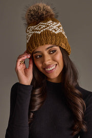 A woman smiles while wearing the Panache Apparel Collection Camel & White Patterned Pom Hat, a brown knit beanie featuring a white zigzag pattern and a faux fur pom-pom, paired with a black long-sleeve top against a plain background.