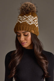 A woman wearing the Panache Apparel Collection Camel & White Patterned Pom Hat, featuring a large fur pom-pom, pairs it with a black ribbed sweater as she looks downward against a neutral background.