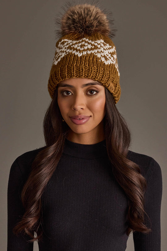 A woman with long, wavy hair wears the Panache Apparel Collection Camel & White Patterned Pom Hat and a black ribbed long-sleeve top, posing against a plain, neutral background.