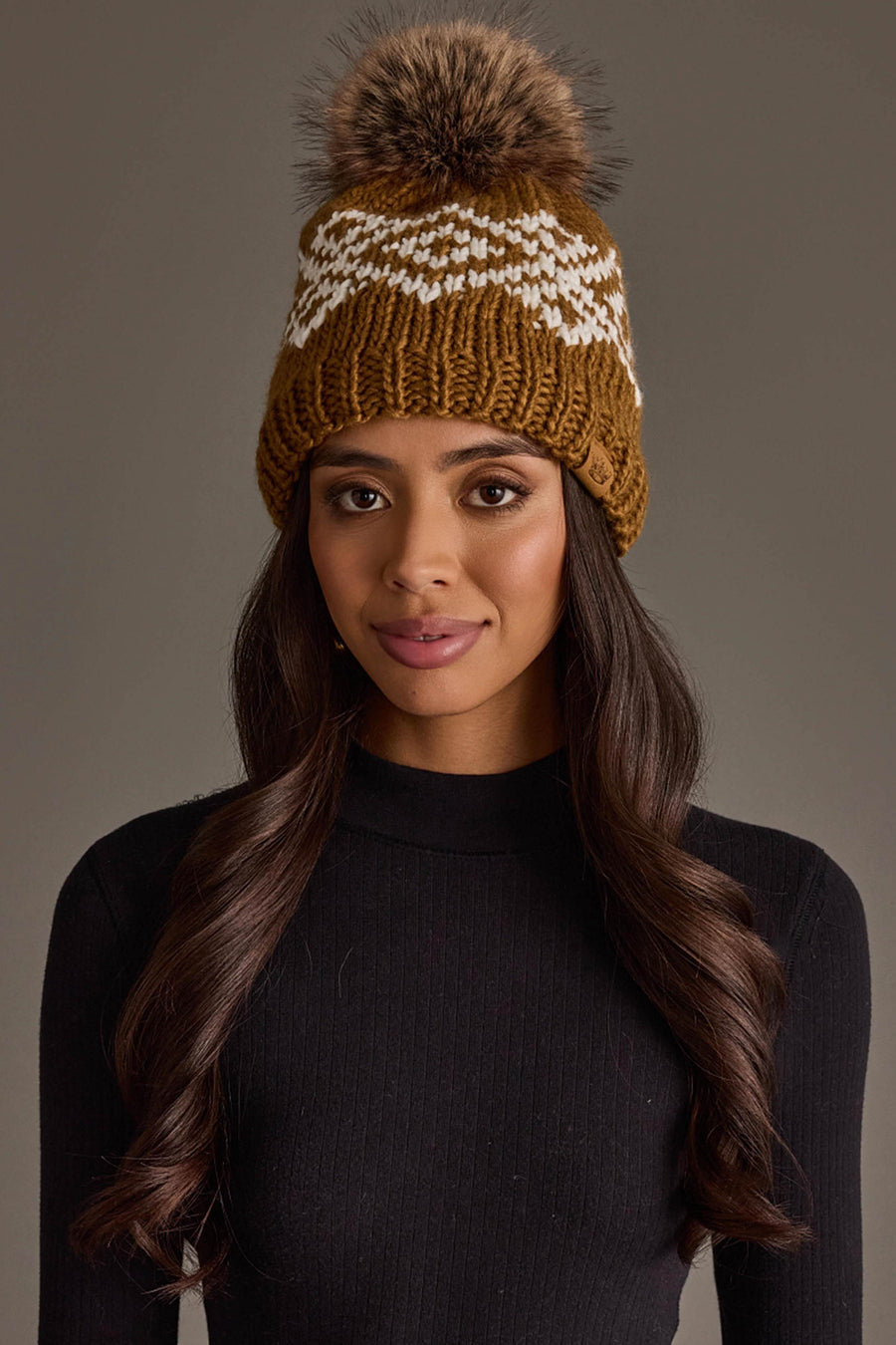 A woman with long, wavy hair wears the Panache Apparel Collection Camel & White Patterned Pom Hat and a black ribbed long-sleeve top, posing against a plain, neutral background.