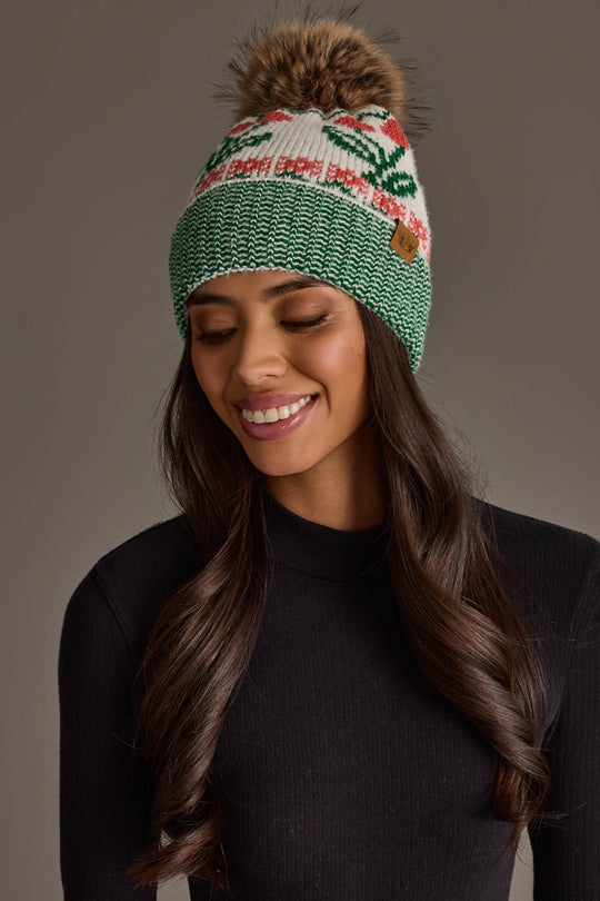 A woman smiles with long wavy hair against a plain gray background, wearing the Panache Apparel Collection Off White Cherry Pattern Pom Hat, featuring green, white, and red cherry details with a faux fur pom-pom, paired with a black ribbed turtleneck sweater.