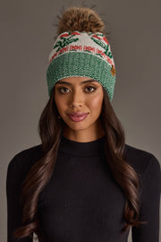 A woman with long dark hair wears the Panache Apparel Collection Off White Cherry Pattern Pom Hat, a green and white knit winter hat featuring a festive cherry design, fleece lining for extra warmth, and a faux fur pom-pom, paired with a black ribbed sweater, standing against a neutral gray background.