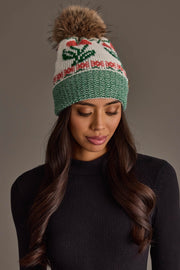 A woman with long, wavy hair and a neutral expression is wearing the Off White Cherry Pattern Pom Hat from Panache Apparel Collection,, a cozy, fleece-lined beanie featuring green trim, a red and green cherry pattern design, and a brown faux fur pom-pom, perfect for adding warmth and style on chilly days.