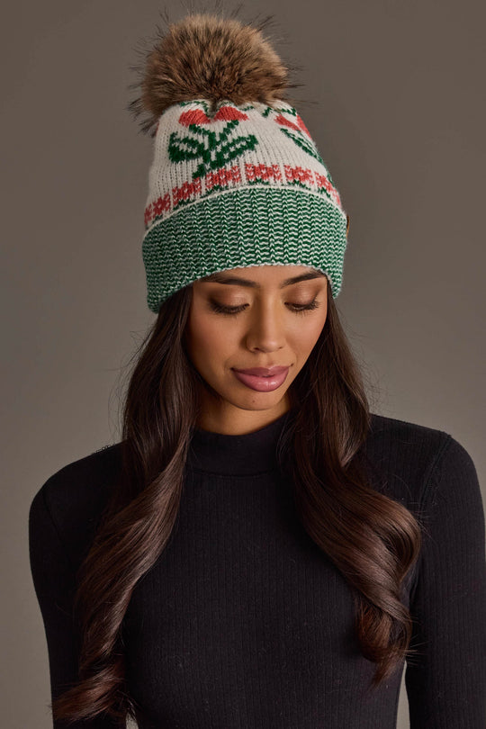 A woman with long, wavy hair and a neutral expression is wearing the Off White Cherry Pattern Pom Hat from Panache Apparel Collection,, a cozy, fleece-lined beanie featuring green trim, a red and green cherry pattern design, and a brown faux fur pom-pom, perfect for adding warmth and style on chilly days.