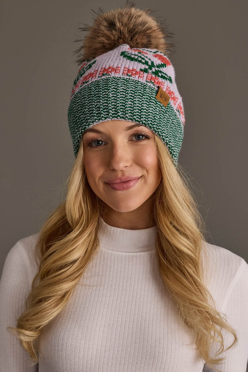 Smiling in front of a plain gray background, a woman with long blonde hair wears the Panache Apparel Collection Light Purple Cherry Pattern Pom Hat and a white ribbed turtleneck.