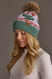 A woman with long blonde hair wears a white sweater and the Light Purple Cherry Pattern Pom Hat by Panache Apparel Collection. The knit winter hat features cherry patterns in light purple along with a large brown pom-pom on top. She is looking down and smiling softly.