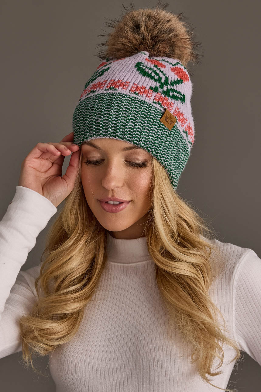 A woman with long blonde hair wears a white ribbed turtleneck and the Panache Apparel Collection Light Purple Cherry Pattern Pom Hat, a knit beanie featuring green, white, and pink cherry designs with a faux fur pom-pom. She is touching the side of her hat and smiling softly.