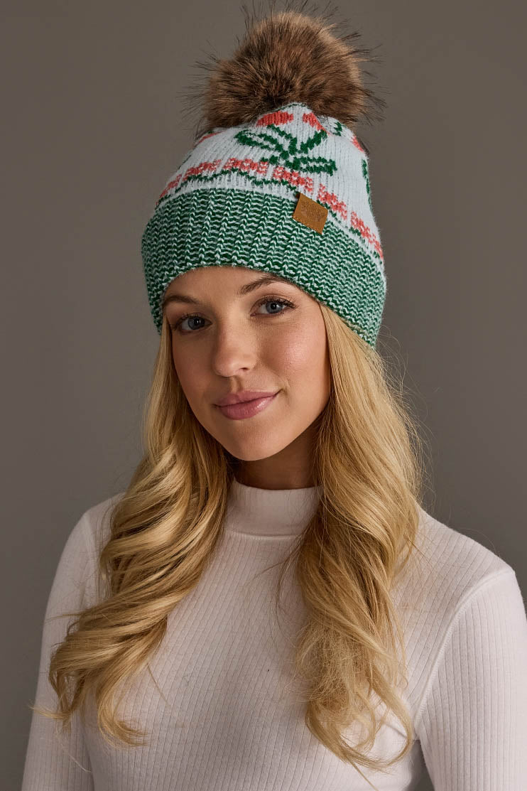 A woman with long blonde hair wears the Panache Apparel Collection Light Blue Cherry Pattern Pom Hat and a white ribbed sweater, standing against a plain gray background.