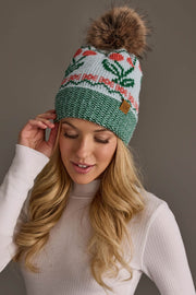 A woman with long blonde hair wears a white shirt and the Light Blue Cherry Pattern Pom Hat by Panache Apparel Collection. She is looking down with a soft smile, gently touching the side of her hat.