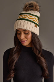 A woman models the Panache Apparel Collection Cream, Green & Gold Patterned Pom Hat—a cozy knit beanie featuring green and yellow zigzag stripes and a faux fur pom-pom—paired with a black long-sleeve top, as she looks down with a soft smile against a plain background.