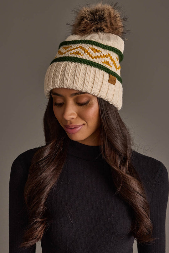 A woman models the Panache Apparel Collection Cream, Green & Gold Patterned Pom Hat—a cozy knit beanie featuring green and yellow zigzag stripes and a faux fur pom-pom—paired with a black long-sleeve top, as she looks down with a soft smile against a plain background.