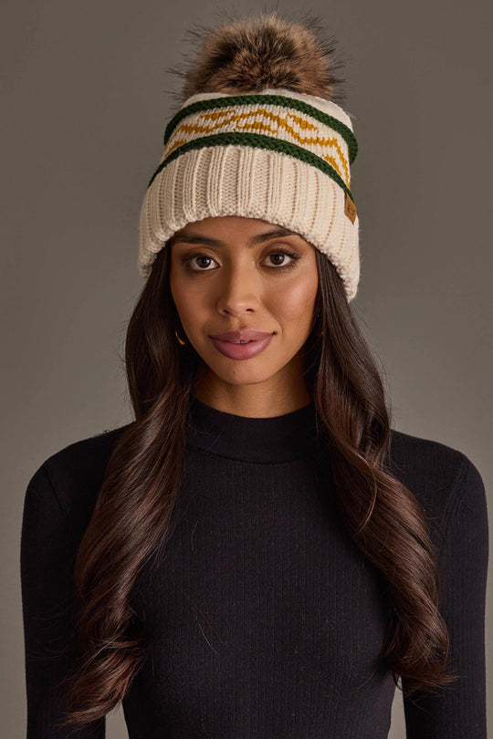 A woman with long brown hair is wearing a black sweater and the Cream, Green & Gold Patterned Pom Hat from Panache Apparel Collection, standing against a plain gray background.