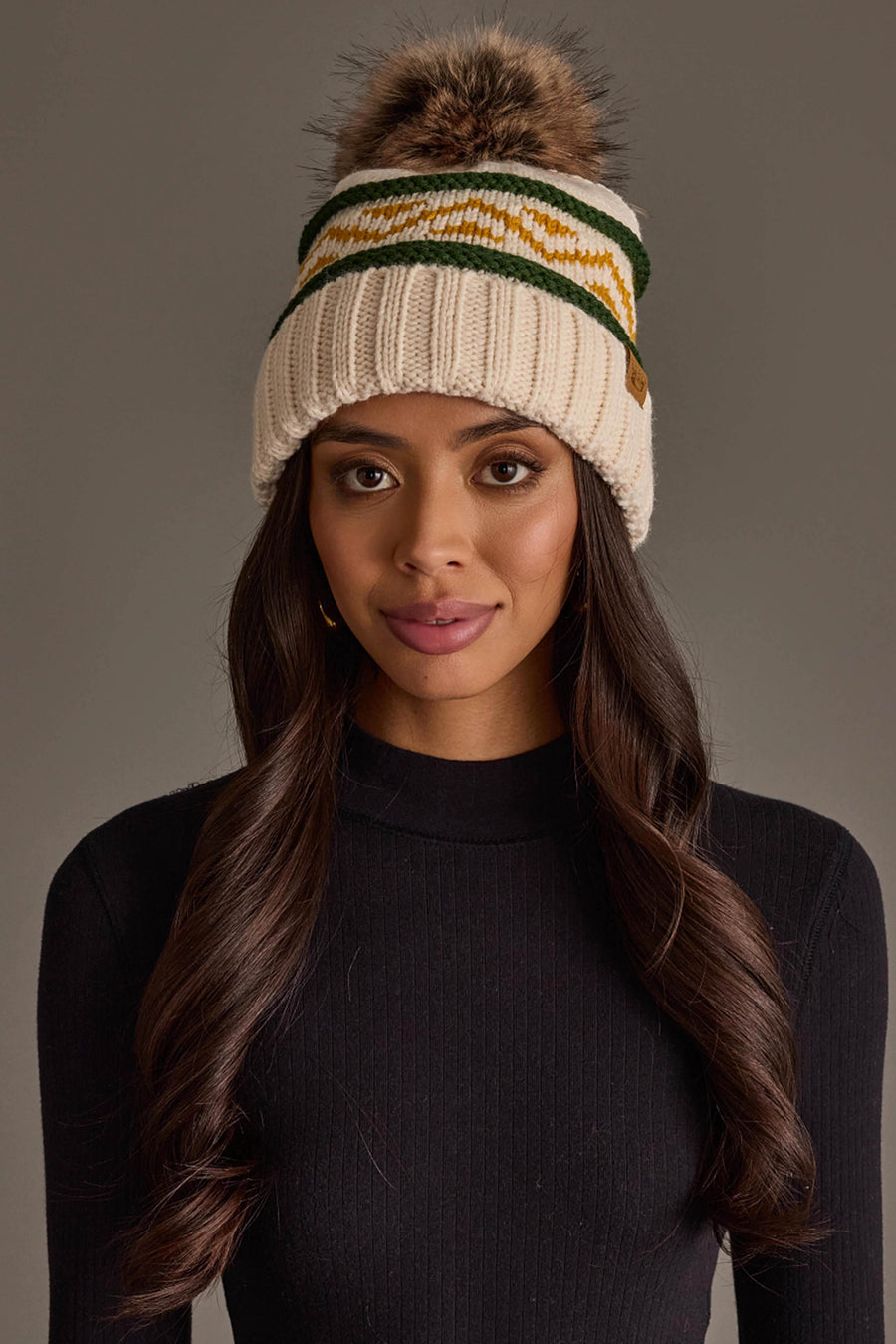 A woman with long brown hair is wearing a black sweater and the Cream, Green & Gold Patterned Pom Hat from Panache Apparel Collection, standing against a plain gray background.