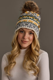 A woman with long blonde hair wears a white ribbed turtleneck and the Panache Apparel Collection’s Ivory, Green, Gray & Gold Patterned Pom Hat, posing against a plain gray background.