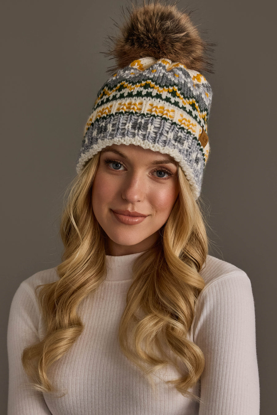 A woman with long blonde hair wears a white ribbed turtleneck and the Panache Apparel Collection’s Ivory, Green, Gray & Gold Patterned Pom Hat, posing against a plain gray background.