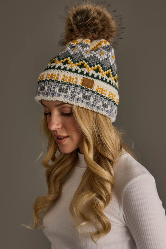 A woman with long blonde hair wears the Panache Apparel Collection Ivory, Green, Gray & Gold Patterned Pom Hat—a knit winter hat featuring a pompom and eye-catching patterns—paired with a white ribbed turtleneck sweater as she looks down against a plain background.