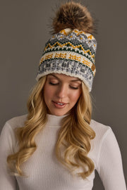 A woman with long blonde hair wears the Panache Apparel Collection Ivory, Green, Gray & Gold Patterned Pom Hat—a knit winter hat topped with a fur pom-pom—along with a white ribbed sweater, smiling softly and looking down against a plain gray background.