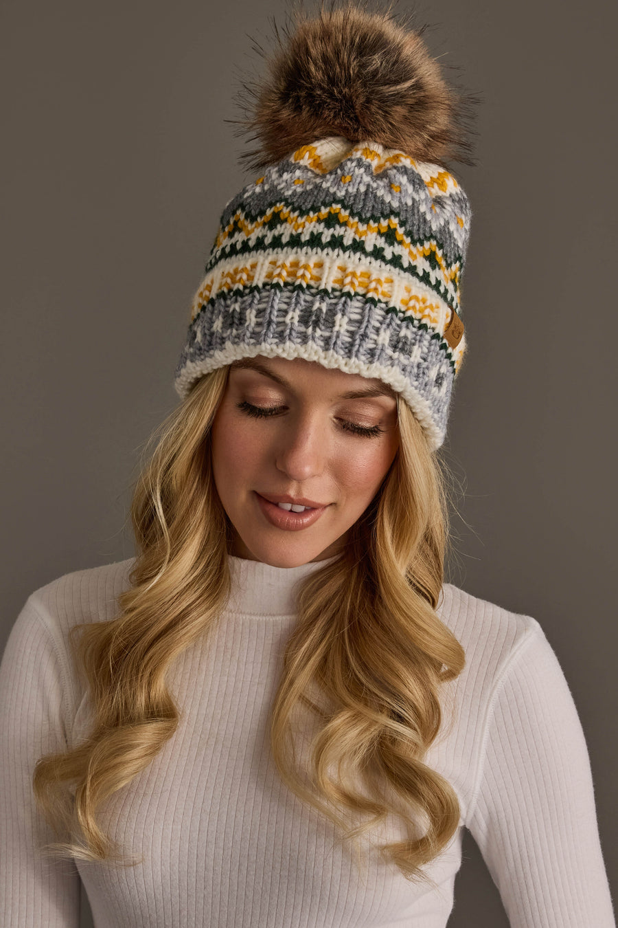 A woman with long blonde hair wears the Panache Apparel Collection Ivory, Green, Gray & Gold Patterned Pom Hat—a knit winter hat topped with a fur pom-pom—along with a white ribbed sweater, smiling softly and looking down against a plain gray background.