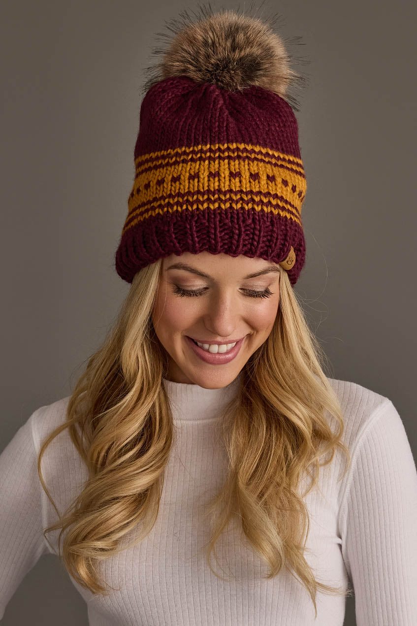 A smiling woman with long blonde hair wears the Maroon & Gold Patterned Knit Pom Hat from Panache Apparel Collection, paired with a white ribbed turtleneck, standing against a plain gray background.