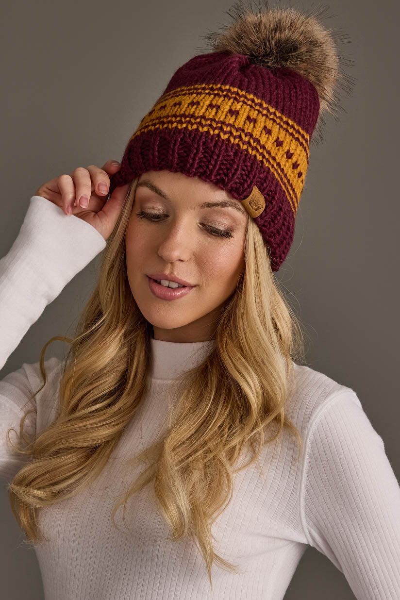 A woman with long blonde hair models the Panache Apparel Collection’s Maroon & Gold Patterned Knit Pom Hat, featuring a fur pom-pom, paired with a white ribbed turtleneck as she poses with one hand touching the hat against a gray background.