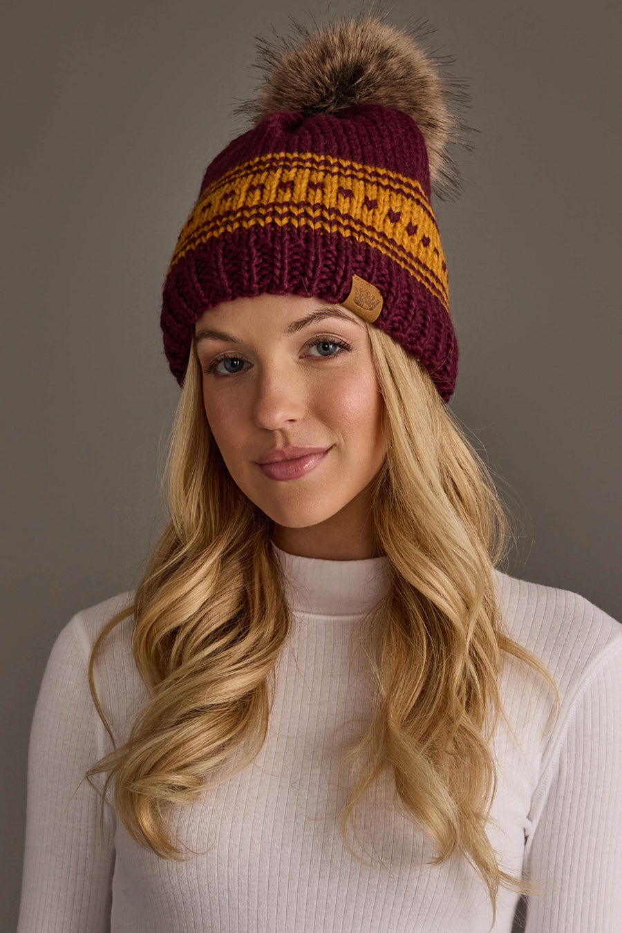 A woman with long blonde hair wears the Maroon & Gold Patterned Knit Pom Hat from Panache Apparel Collection and a white ribbed turtleneck, smiling softly against a plain gray background.