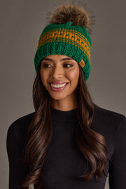 A woman with long brown hair is smiling against a plain gray background while wearing the Panache Apparel Collection’s Green & Gold Patterned Knit Pom Hat and a black ribbed sweater.
