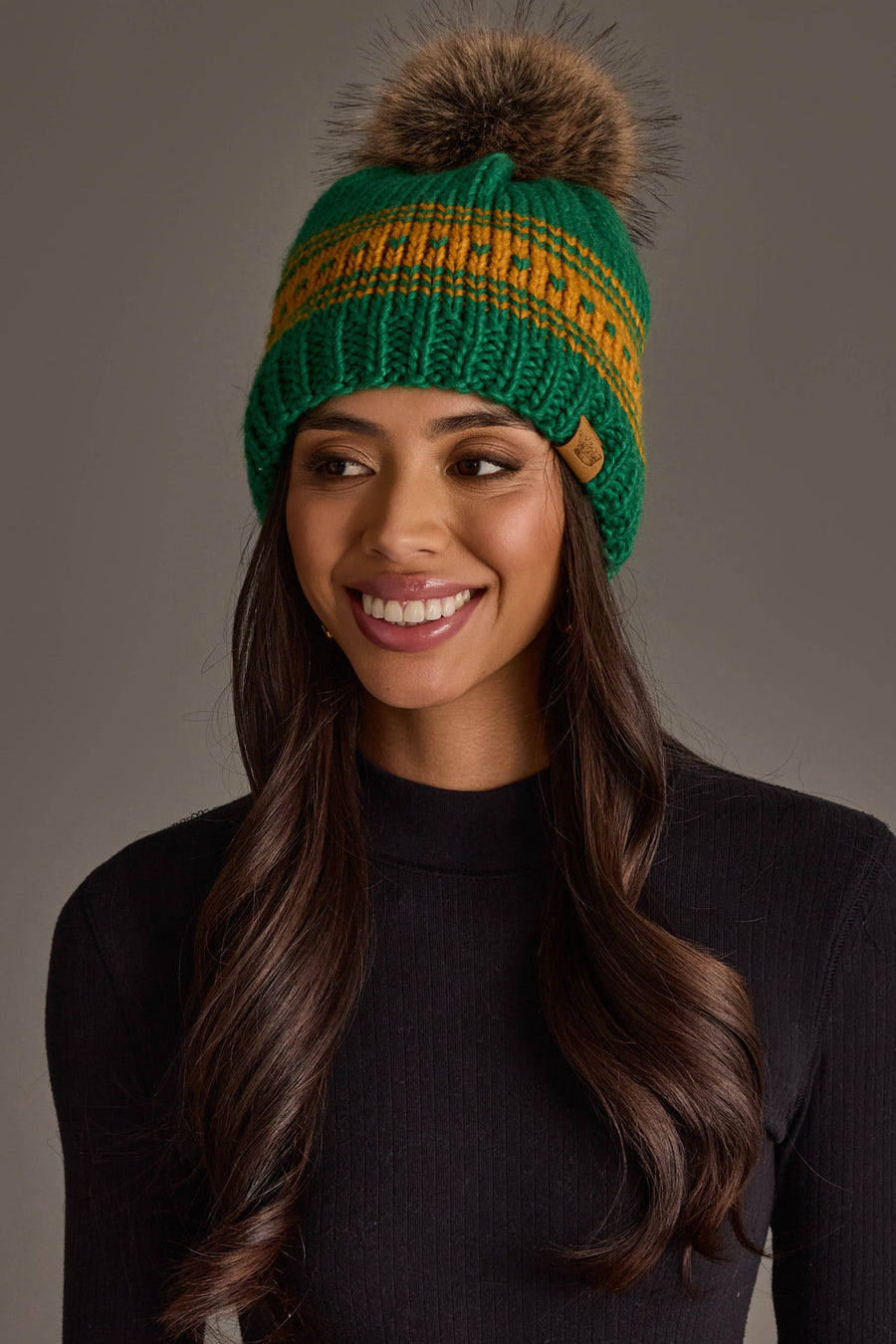 A woman with long brown hair is smiling against a plain gray background while wearing the Panache Apparel Collection’s Green & Gold Patterned Knit Pom Hat and a black ribbed sweater.