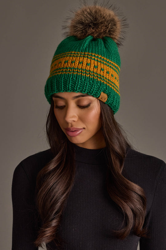 A woman with long brown hair, wearing the Panache Apparel Collection Green & Gold Patterned Knit Pom Hat featuring a brown pom-pom and paired with a black turtleneck sweater, looks down against a plain background.