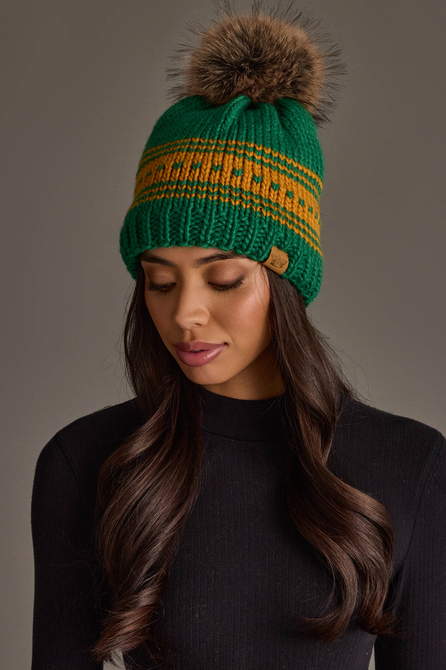 A woman with long brown hair, wearing the Panache Apparel Collection Green & Gold Patterned Knit Pom Hat featuring a brown pom-pom and paired with a black turtleneck sweater, looks down against a plain background.