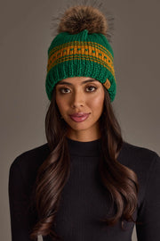 A woman with long brown hair and a black ribbed top models the Panache Apparel Collection's Green & Gold Patterned Knit Pom Hat, featuring a vibrant yellow stripe and fur pom-pom, against a plain gray background.