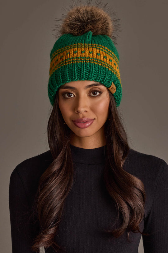 A woman with long brown hair and a black ribbed top models the Panache Apparel Collection's Green & Gold Patterned Knit Pom Hat, featuring a vibrant yellow stripe and fur pom-pom, against a plain gray background.