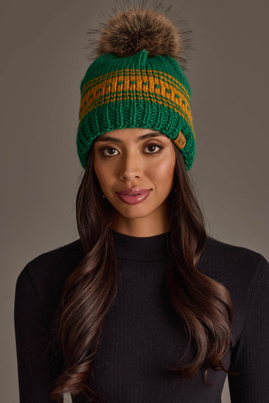 A woman with long brown hair and a black ribbed top models the Panache Apparel Collection's Green & Gold Patterned Knit Pom Hat, featuring a vibrant yellow stripe and fur pom-pom, against a plain gray background.