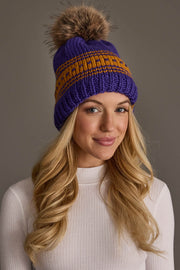 A woman with long blonde hair is wearing a white ribbed top and the Panache Apparel Collection Purple & Gold Patterned Knit Pom Hat, featuring a brown faux fur pom-pom, while standing against a plain gray background.