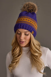 A woman with long blonde hair models the Panache Apparel Collection Purple & Gold Patterned Knit Pom Hat, featuring a brown fur pom-pom, paired with a white ribbed turtleneck top against a plain gray background.
