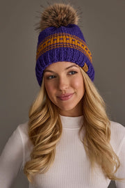 A woman with long blonde hair smiles softly against a plain gray background, wearing the Panache Apparel Collection's Purple & Gold Patterned Knit Pom Hat and a white ribbed long-sleeve top.